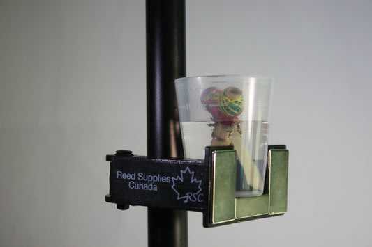 A reed soaker and tool holder mounted on a black metal rod, with a clear plastic water cup containing a reed, and the label 'Reed Supplies Canada'.